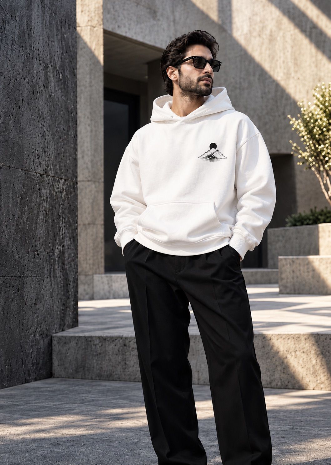 A man in sunglasses wearing a white hoodie with a small mountain print, in a stone courtyard